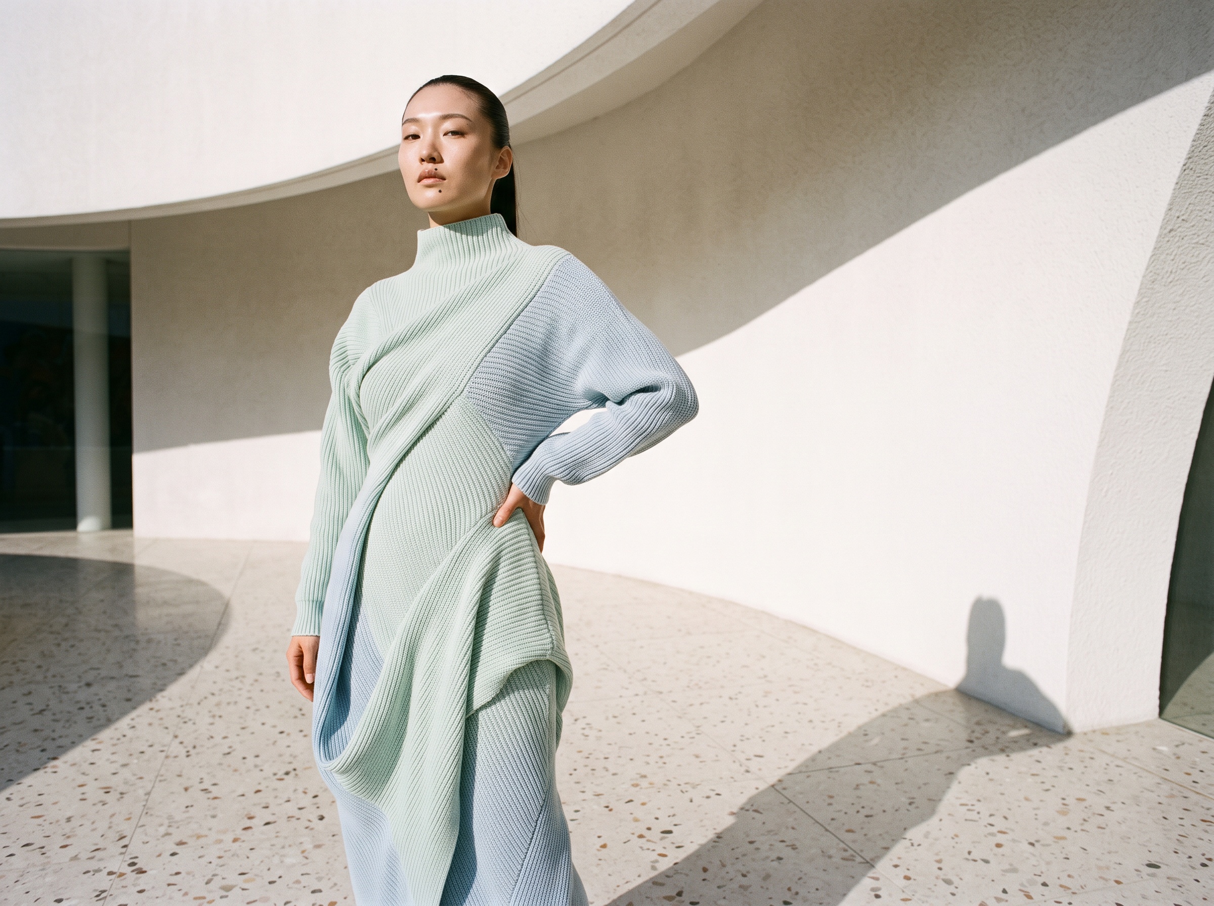 An AI fashion model in a seamless knitted dress in pale mint and powder blue, one continuous sculpted piece, in a bright white gallery
